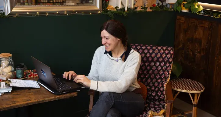 Anna Grau-Galofré, souriante, en pull clair, travaille sur un ordinateur portable dans un café cosy. Assise sur un fauteuil à motifs, elle semble détendue, une tasse de café posée sur la table en bois.