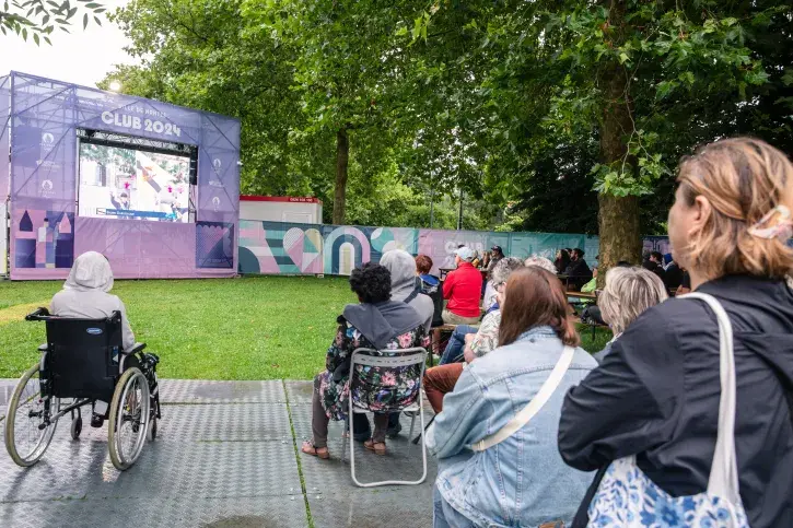 Du public assis, et un personne en fauteuil roulant, sur une plateforme en dur, devant la pelouse sur laquelle est posée le grand écran diffusant les jeux olympiques