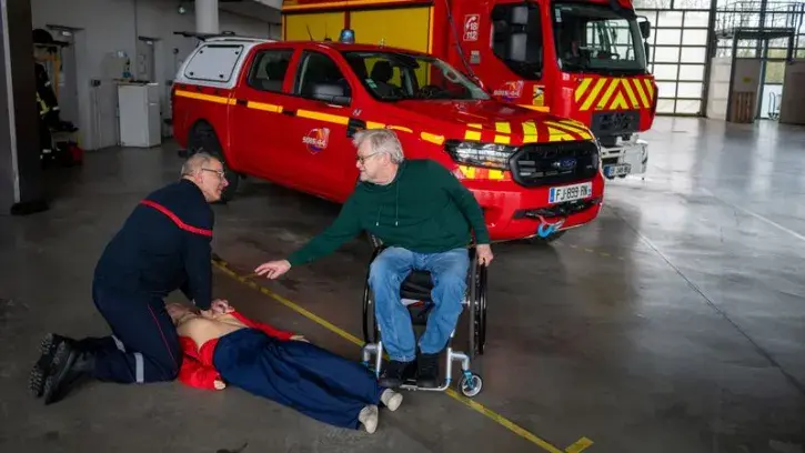 Photographie d'une personne en fauteuil roulant indiquant la marche à suivre sur les premiers gestes de secours à une autre personne, effectuant un massage cardiaque sur un mannequin d'entrainement, le tout dans une caserne de pompier.